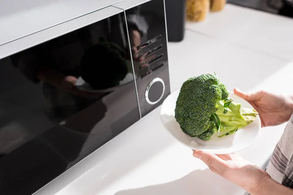 Cropped view of woman holding plate with broccoli near microwave — Stock Photo