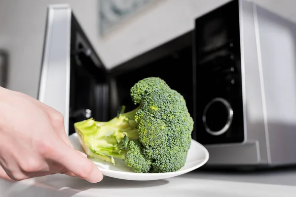 Vista ritagliata della donna che tiene il piatto con broccoli vicino al forno a microonde — Foto stock