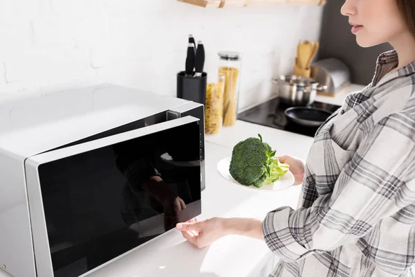 Cropped view of woman holding plate with broccoli and opening microwave — Stock Photo