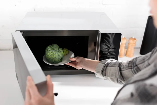Vista ritagliata della donna che tiene il piatto con broccoli e lo mette nel forno a microonde — Foto stock