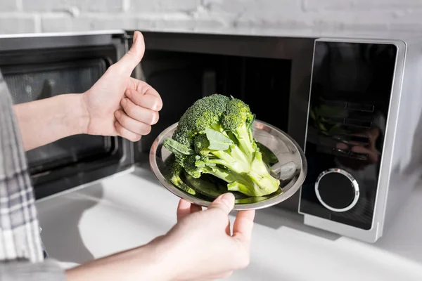 Vue recadrée de la femme tenant plaque avec brocoli près de micro-ondes et montrant comme — Photo de stock