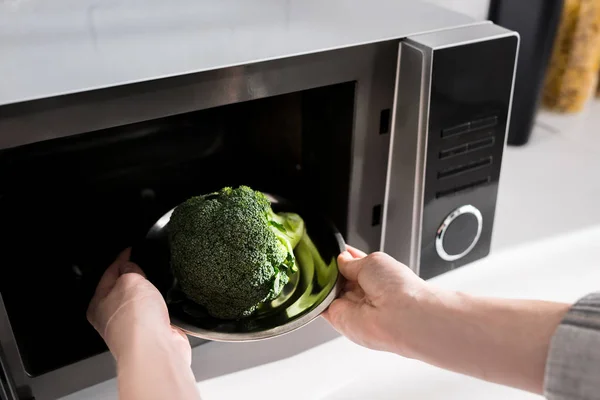 Vista ritagliata della donna che tiene il piatto con broccoli e lo mette nel forno a microonde — Foto stock