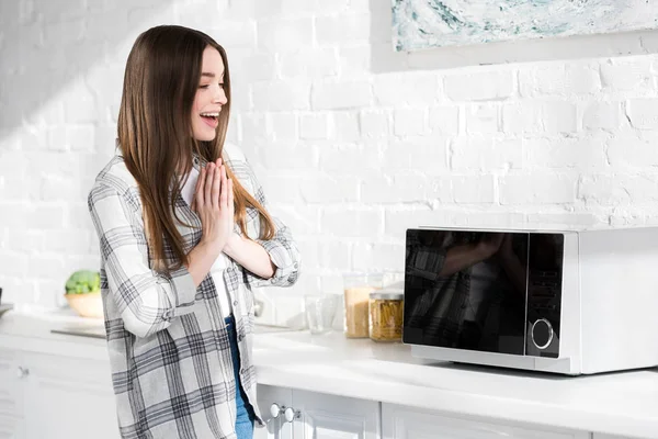 Mujer sonriente y atractiva con las manos orantes mirando al microondas en la cocina - foto de stock