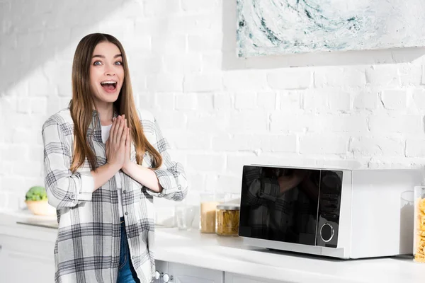 Shocked and attractive woman with praying hands standing near microwave in kitchen — Stock Photo