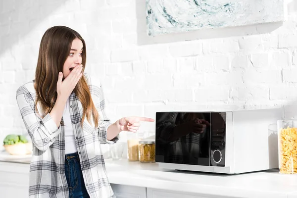 Schockierte und attraktive Frau zeigt mit dem Finger auf Mikrowelle in Küche — Stockfoto