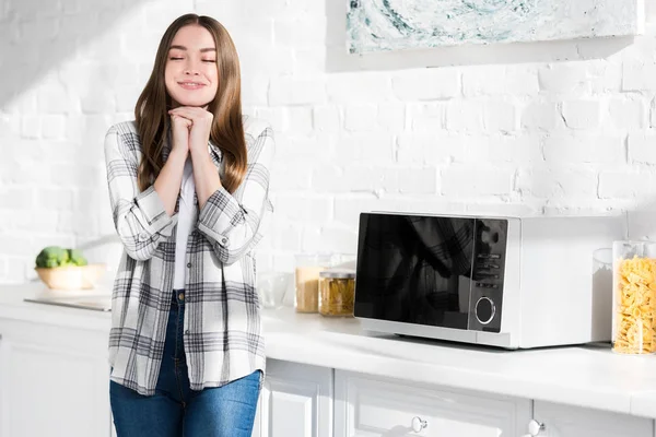 Mujer sonriente y atractiva con los ojos cerrados de pie cerca de microondas en la cocina - foto de stock