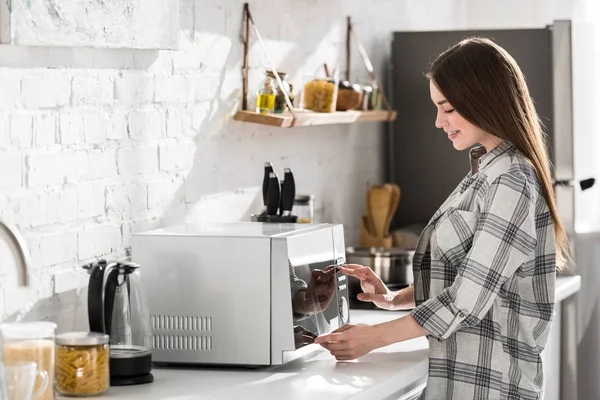 Вид сбоку улыбающейся женщины в рубашке, использующей микроволновку на кухне — стоковое фото