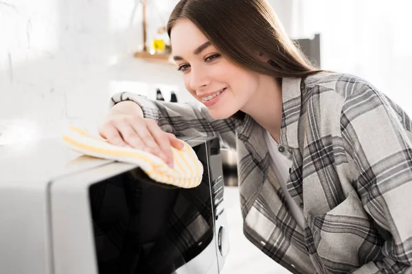 Donna sorridente e attraente pulizia microonde con straccio in cucina — Foto stock