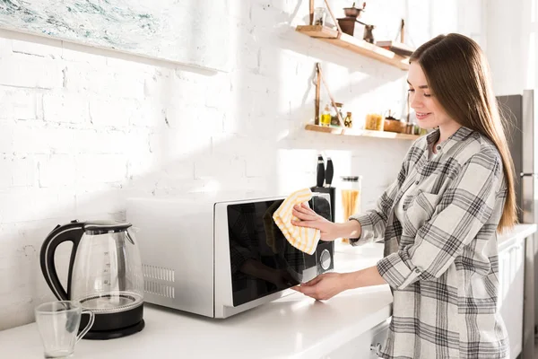 Mulher sorridente e atraente limpeza de microondas com pano na cozinha — Fotografia de Stock