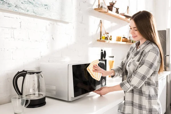 Femme souriante et attrayante nettoyage micro-ondes avec chiffon dans la cuisine — Photo de stock