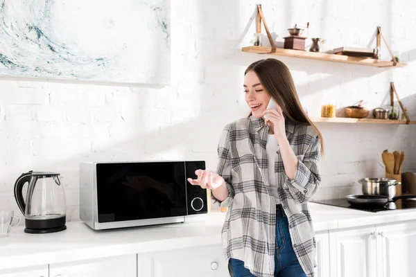 Mujer sonriente hablando en teléfono inteligente cerca de microondas en la cocina - foto de stock