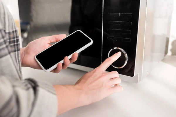 Cropped view of woman holding smartphone and using microwave in kitchen — Stock Photo