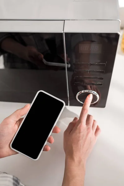 Cropped view of woman holding smartphone and using microwave in kitchen — Stock Photo