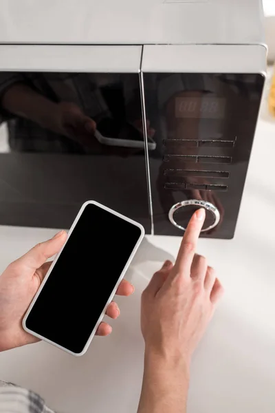 Vista recortada de la mujer sosteniendo teléfono inteligente y el uso de microondas en la cocina — Stock Photo