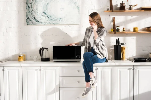 Smiling woman talking on smartphone near microwave in kitchen — Stock Photo