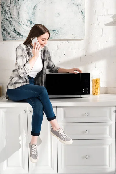 Mujer sonriente hablando en el teléfono inteligente y mirando al microondas en la cocina - foto de stock