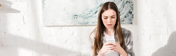 Panoramic shot of shocked woman using smartphone in kitchen — Stock Photo