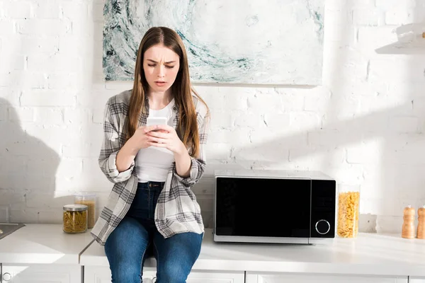 Шокированная женщина с помощью смартфона рядом с микроволновой печью на кухне — стоковое фото