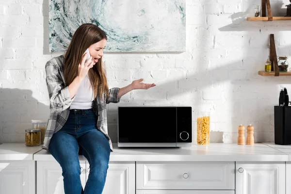 Mujer enojada hablando en el teléfono inteligente y mirando microondas en la cocina - foto de stock