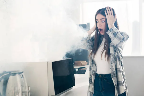 Schockierte und attraktive Frau blickt auf kaputte Mikrowelle in Küche — Stockfoto