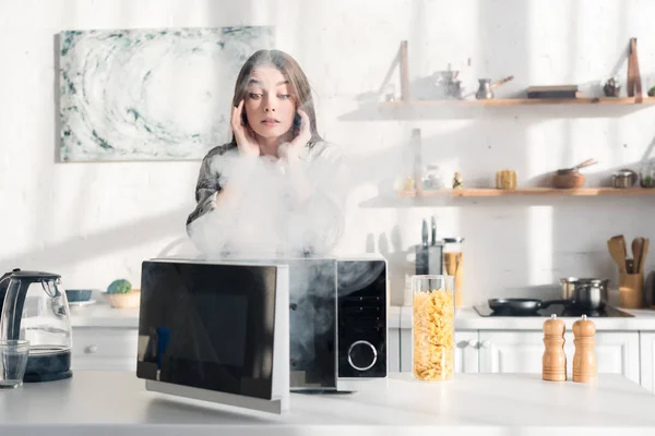 Femme choquée et attrayante regardant micro-ondes cassé dans la cuisine — Photo de stock