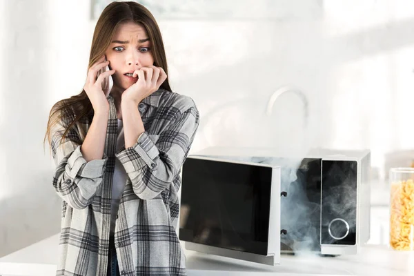 Scared woman talking on smartphone near broken microwave in kitchen — Stock Photo