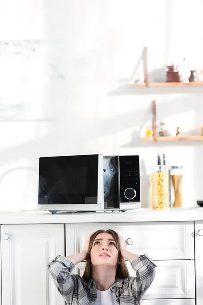 Traurige und attraktive Frau blickt auf kaputte Mikrowelle in Küche — Stockfoto