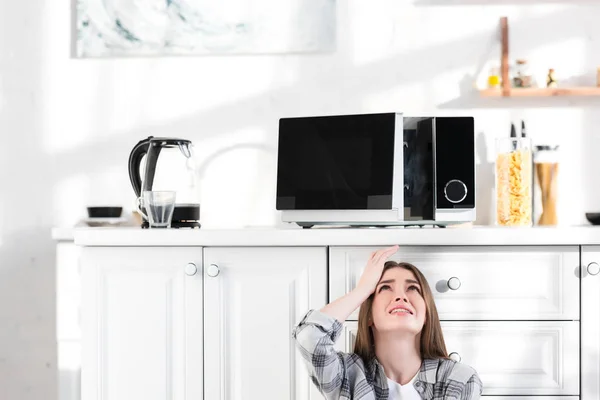 Traurige und attraktive Frau blickt auf kaputte Mikrowelle in Küche — Stockfoto