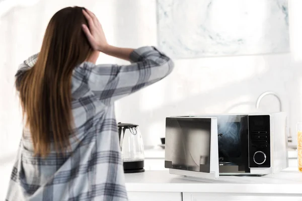 Visão traseira da mulher chocada olhando para o microondas quebrado na cozinha — Fotografia de Stock