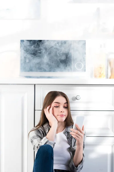 Sad woman using smartphone near broken microwave in kitchen — Stock Photo