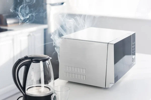 Hervidor eléctrico, microondas roto y humeante en la mesa en la cocina - foto de stock