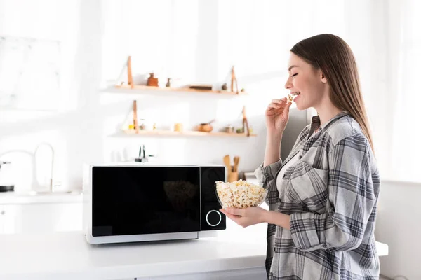 Side view of smiling and attractive woman eating popcorn near microwave — Stock Photo