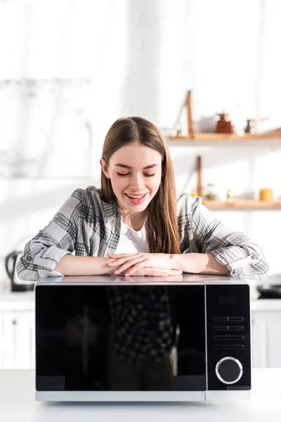 Улыбающаяся и привлекательная женщина смотрит на микроволновку на кухне — стоковое фото