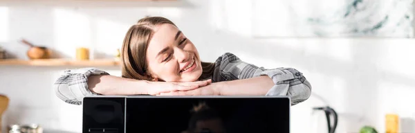 Panoramic shot of smiling and attractive woman lying on microwave in kitchen — Stock Photo