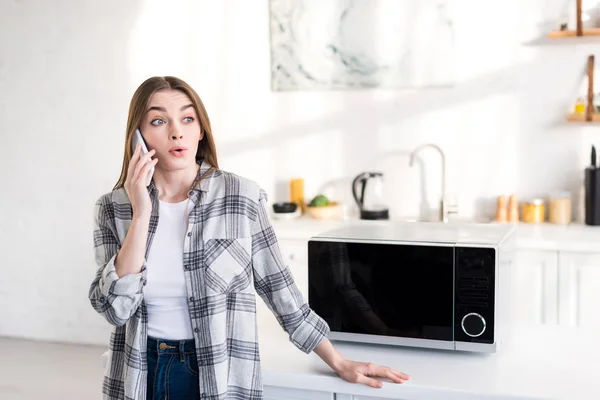 Mulher chocada falando no smartphone perto de microondas na cozinha — Fotografia de Stock