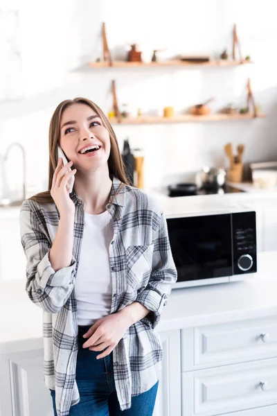 Mujer sonriente hablando en teléfono inteligente cerca de microondas en la cocina - foto de stock