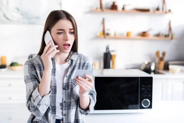 Шокована жінка говорить на смартфоні біля мікрохвильовки і тримає візитну картку на кухні — стокове фото