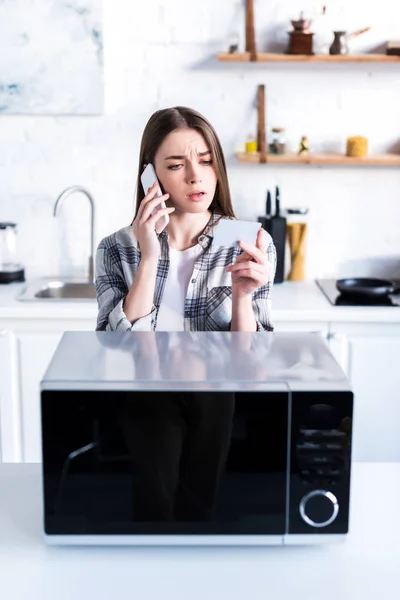 Traurige Frau spricht auf Smartphone in der Nähe der Mikrowelle und hält Visitenkarte in der Küche — Stockfoto