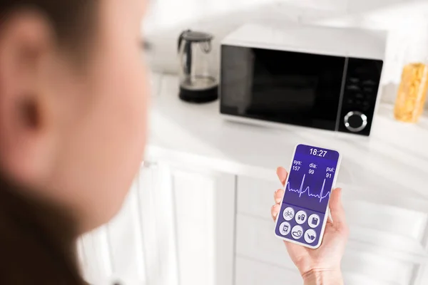 Selective focus of woman holding smartphone with health app and microwave on background — Stock Photo