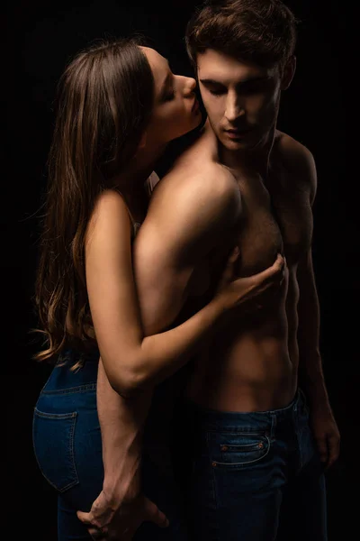 Привлекательная молодая женщина, обнимающая и целующая сексуального парня, изолированного от черного — стоковое фото