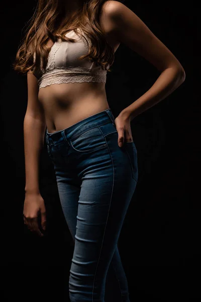 Обрезанный вид сексуальной молодой девушки в бюстгальтере и джинсах позируя с рукой в кармане изолированы на черном — стоковое фото
