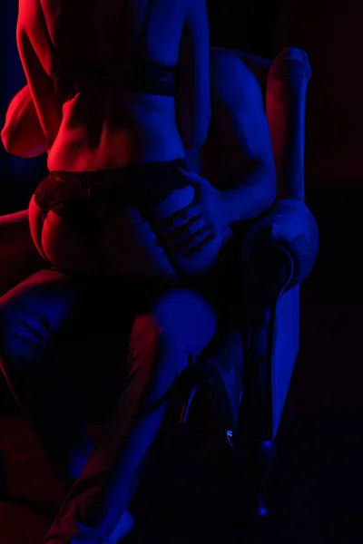 Vue recadrée de femme sexy en sous-vêtements assis sur le petit ami dans le fauteuil dans la lumière rouge isolé sur noir — Photo de stock