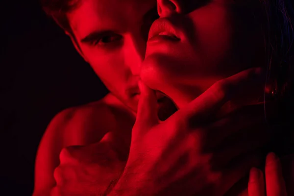 Desnudo apasionado hombre sosteniendo cuello de novia en rojo luz aislado en negro - foto de stock