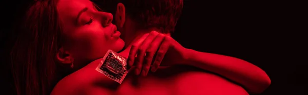 Passionate undressed sexy young couple hugging with condom in red light isolated on black, panoramic shot — Stock Photo