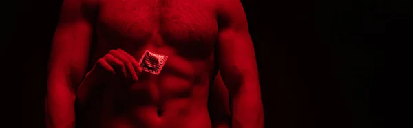 Cropped view of woman touching bare torso of sexy man and holding condom in red light isolated on black, panoramic shot — Stock Photo