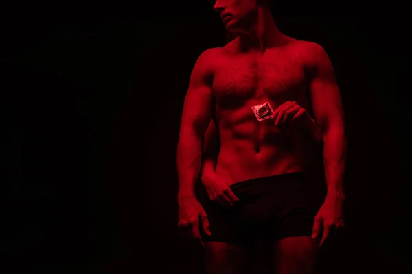 Vue recadrée de la femme touchant torse nu de l'homme sexy et tenant préservatif dans la lumière rouge isolé sur noir — Photo de stock