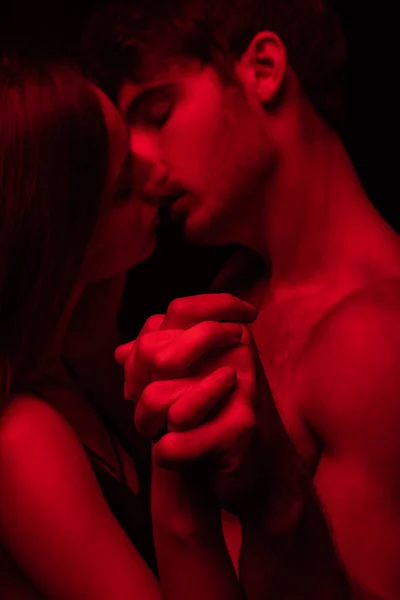 Vue latérale de passionné déshabillé jeune couple sexy embrasser et tenir la main dans la lumière rouge isolé sur noir — Photo de stock