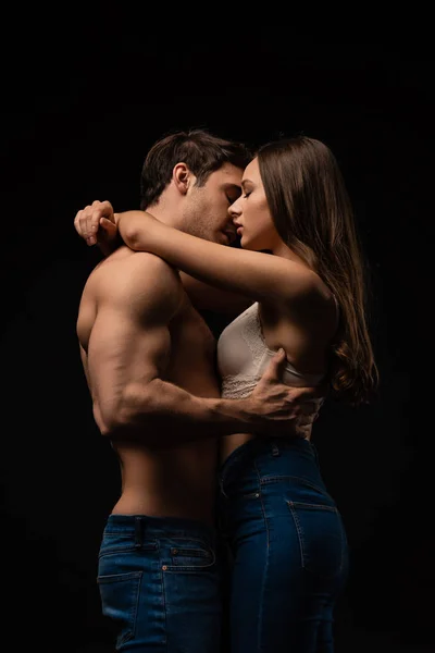 Vista lateral de vestido sexy jovem casal em jeans abraçando e beijando isolado no preto — Fotografia de Stock