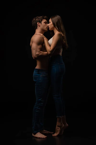 Vista lateral de pareja joven sexy desnuda apasionada en jeans abrazos y besos aislados en negro - foto de stock