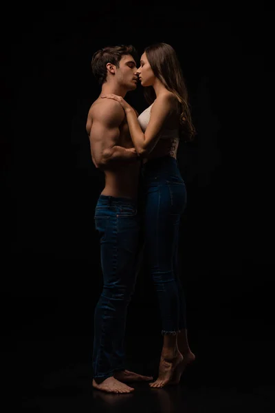 Vista lateral de pareja joven sexy desnuda apasionada en jeans abrazos y besos aislados en negro - foto de stock